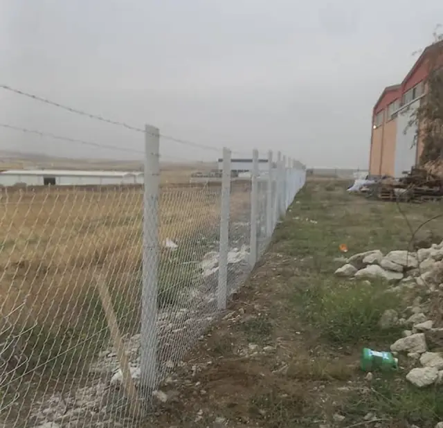 Boş arazi kenarında beton direkler arasında gerdirilmiş tel örgü ve dikenli tel